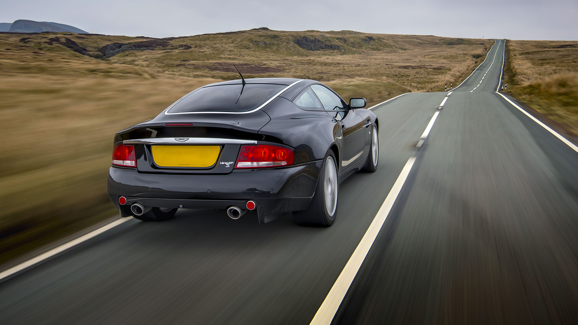 Rückansicht eines schnittigen schwarzen Aston Martin Vanquish S, der auf einer malerischen Landstraße fährt. 