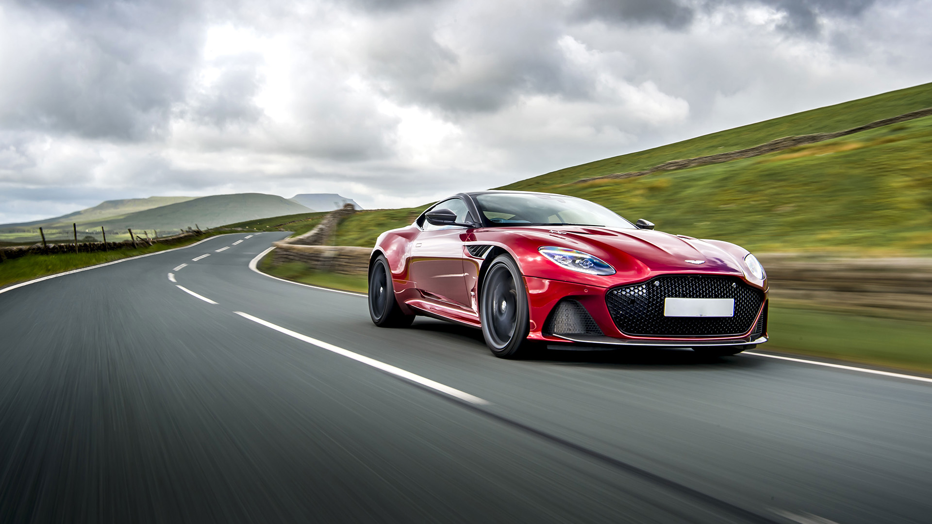 Roter Aston Martin DBS fährt schnell auf einer Landstraße mit grauem Himmel darüber.