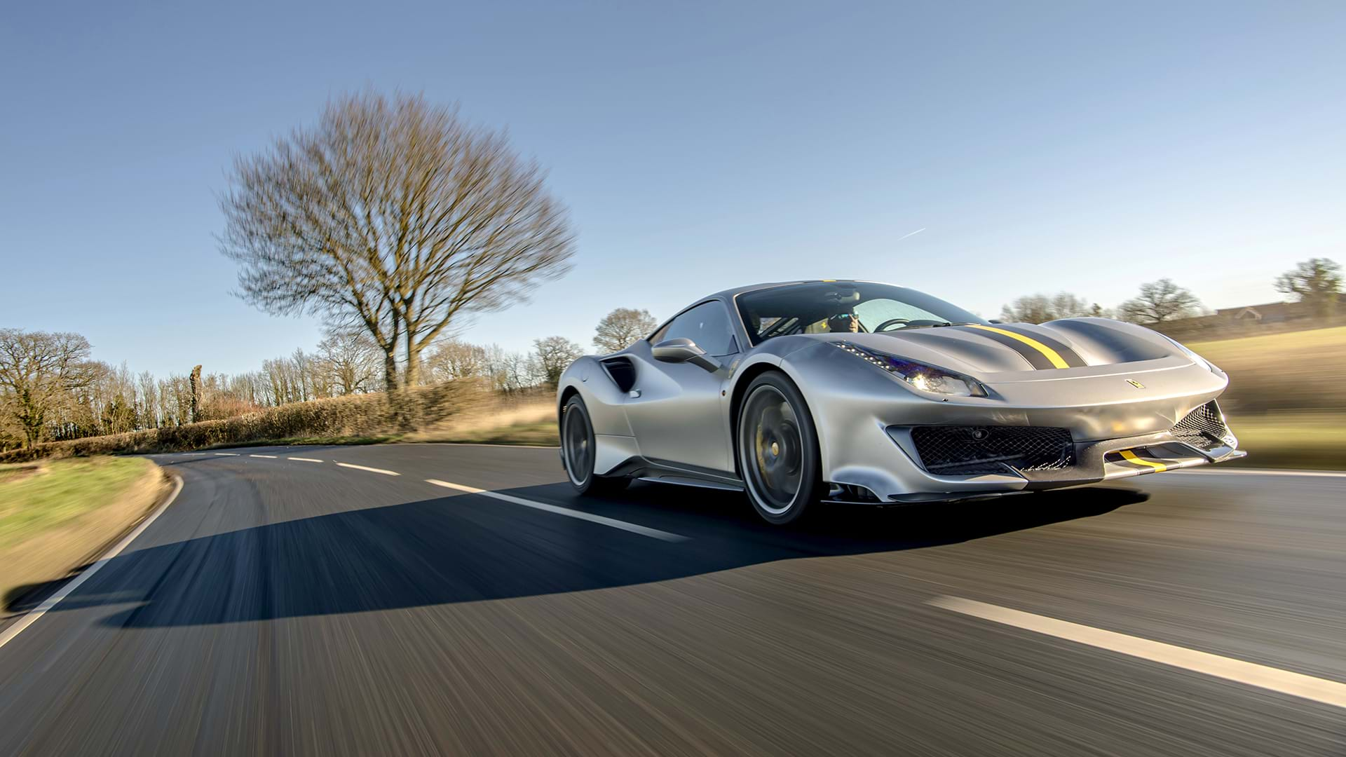 Ein eleganter grauer Ferrari 488 Pista mit einem auffälligen Rennstreifen fährt durch eine malerische Landschaft. 