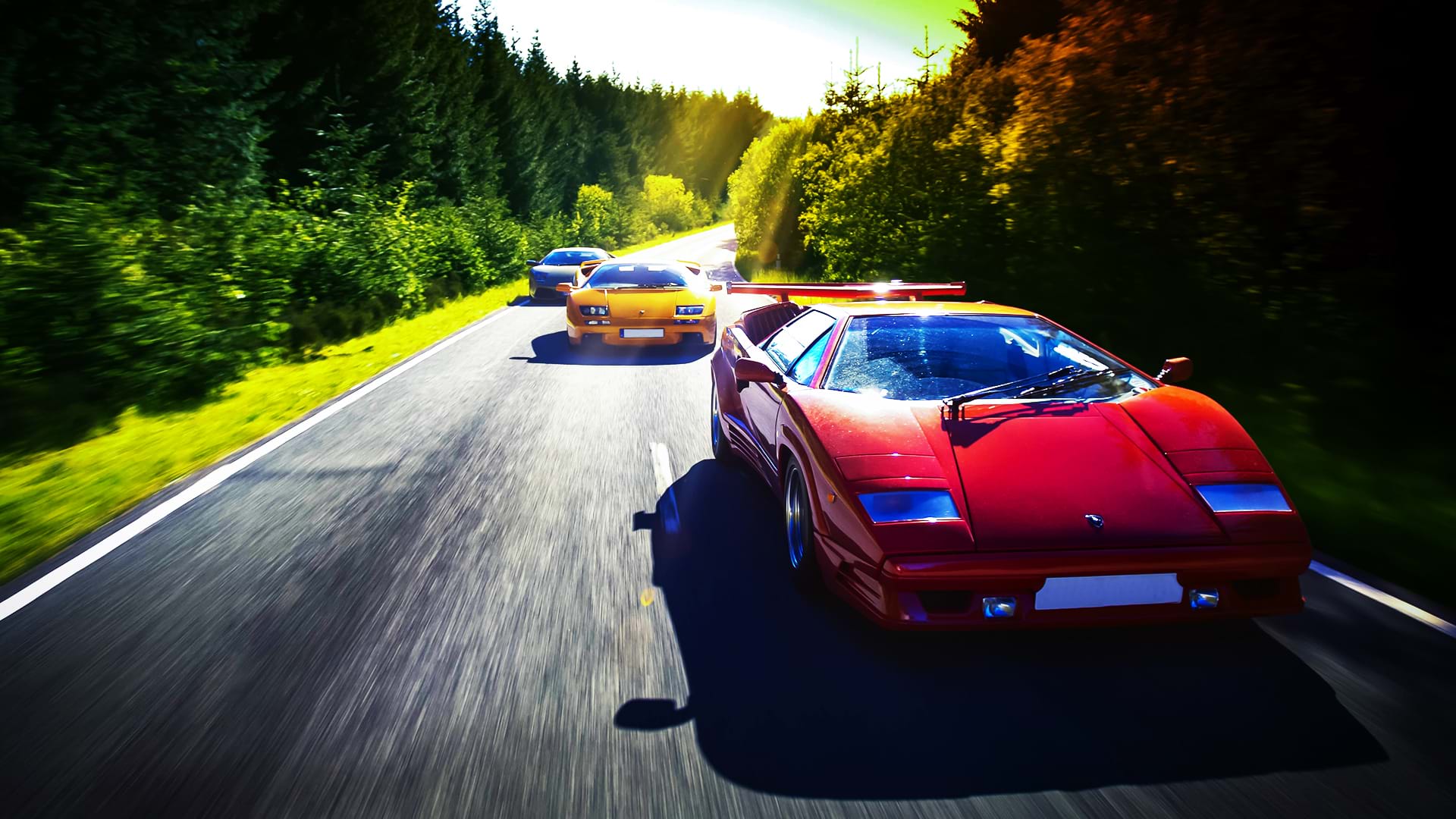 Ein farbenfroher Lamborghini-Konvoi: Countach 25th Anniversary, Diablo 6.0 und Murciélago rast an einem hellen Tag eine Panoramastraße entlang.