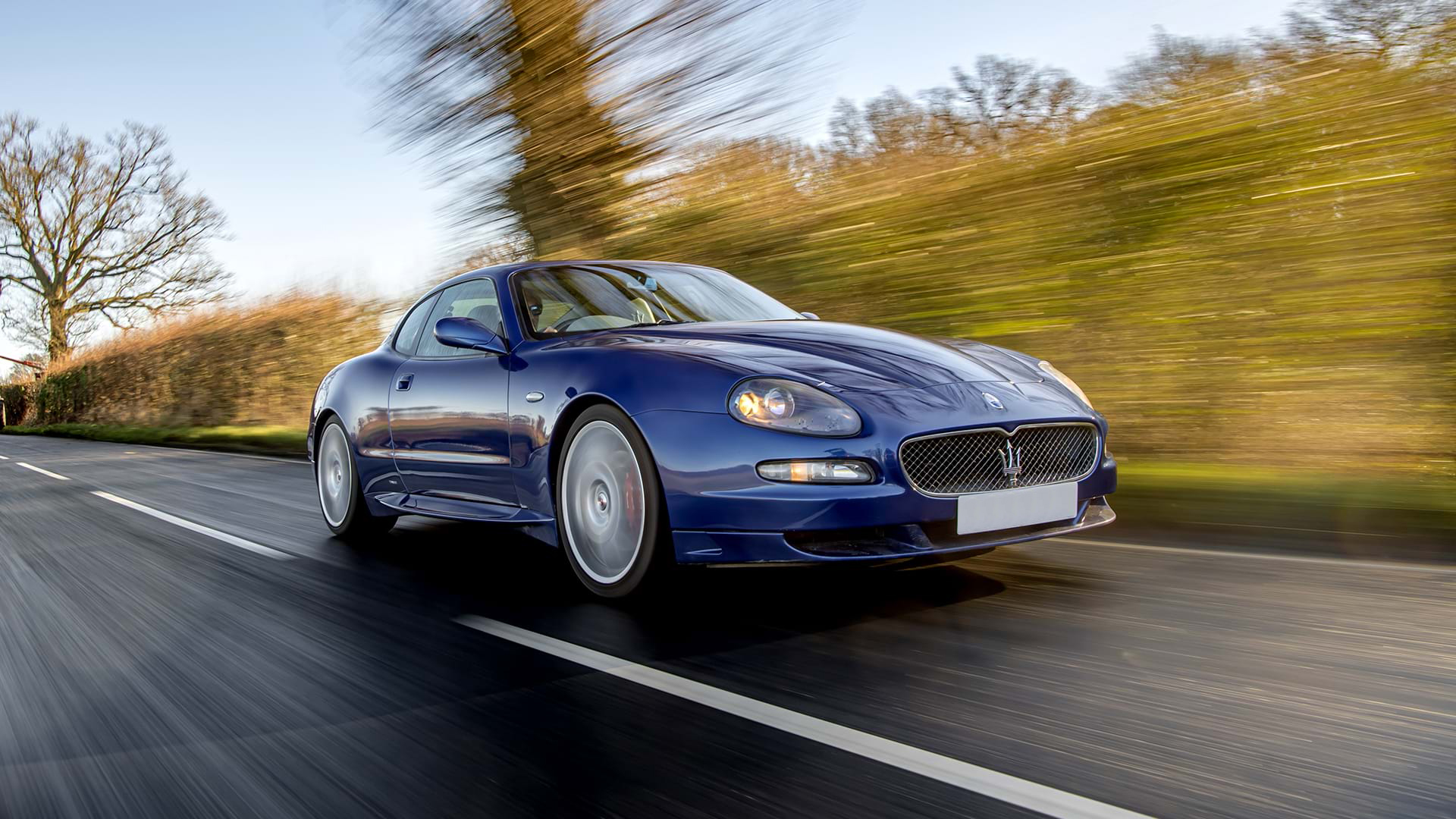 Klassischer blauer Maserati GranSport fährt auf einer Landstraße unter blauem Himmel.