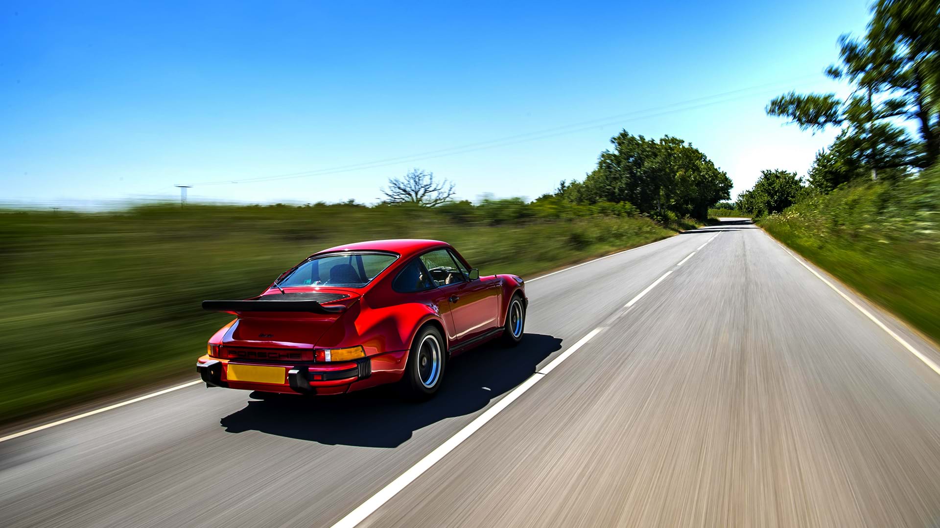 Ein roter Vintage-Turbo Porsche 930 braust an einem sonnigen Tag über eine schöne Landstraße.